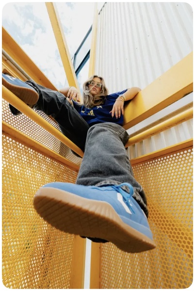 A vertical 2:3 aspect ratio ad showing a close-up, bottom-up view of a blue sneaker on a person, emphasizing the product profile against a bright, architectural backdrop.