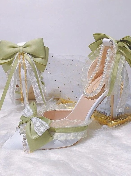 A close-up studio shot of white wedding heels featuring sage green ribbons, lace trim, and pearl strand detailing.