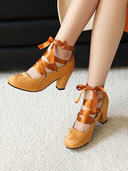 A close-up product shot featuring ochre-toned velvet lace-up block heels, highlighting the delicate ribbon detail and texture.