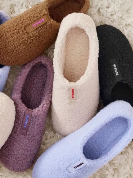 A top-down, high-angle lifestyle shot featuring a collection of fuzzy slippers in various muted colors arranged on a plush white surface.