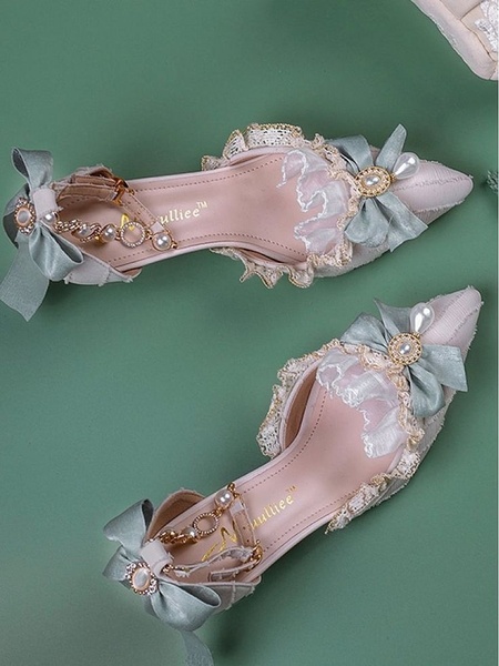A high-angle, top-down product shot of a pair of pastel lace-trimmed bow heels, set against a solid muted green background to emphasize product design.