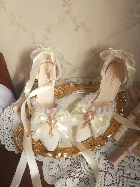 A delicate pair of cream-colored heels adorned with ribbons and pearls, staged on a vintage gold mirror to emphasize a romantic, high-fashion aesthetic.