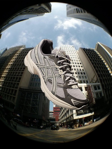A low-angle fisheye lens shot of a single running shoe suspended against a city skyline, emphasizing scale and urban grit.