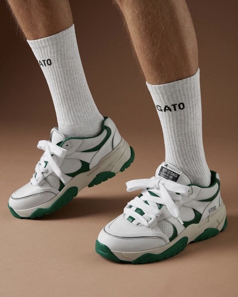 A high-quality product shot of white and green sneakers worn with branded socks against a neutral, earth-toned background.