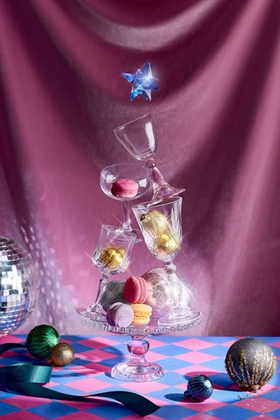 A surreal product shot featuring a precarious, floating stack of champagne flutes filled with macarons and ornaments against a draped velvet backdrop.