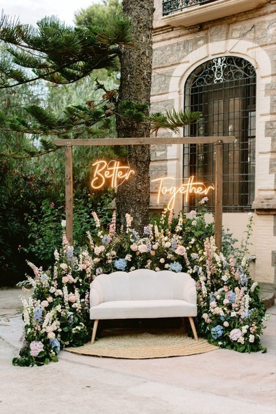 A high-end, romantic wedding lounge area styled with a 'Better Together' neon sign, a velvet sofa, and a lush floral arrangement.