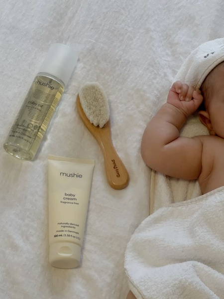 A high-angle top-down shot of baby care products including oil, cream, and a soft-bristled brush, arranged neatly on a textured white bed sheet next to a baby.