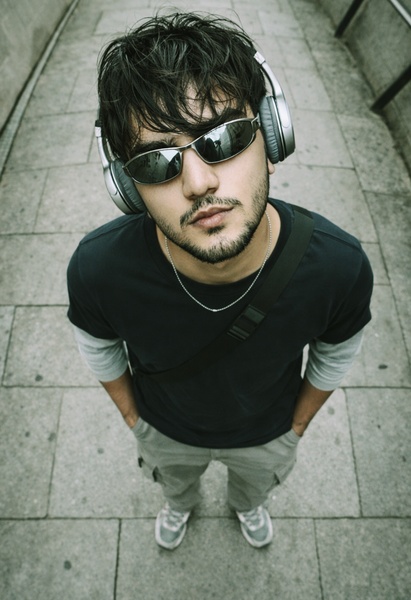 A close-up, high-angle portrait of a man wearing wireless headphones and dark sunglasses in an urban setting.