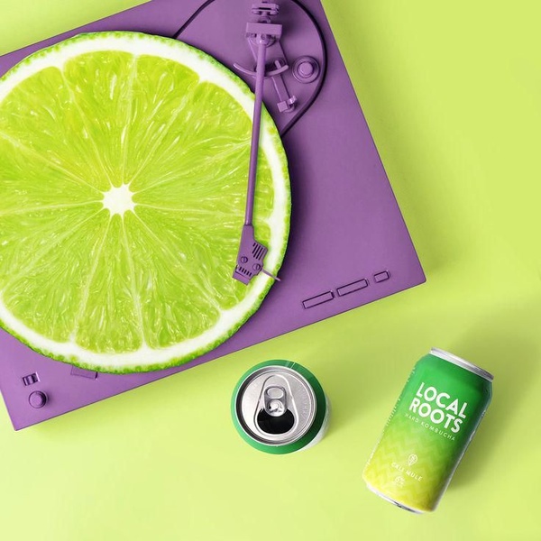 A top-down, square format still life featuring a bright green lime slice placed on a purple record player, paired with a matching hard kombucha can.