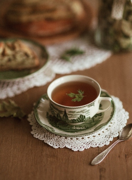 A high-quality lifestyle shot featuring a decorative tea cup on a doily, set against a warm, rustic backdrop with a slice of cake.