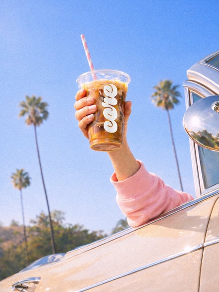 A high-energy, sun-drenched lifestyle shot showing a hand holding an iced coffee drink out of a car window, set against palm trees and a bright blue sky.