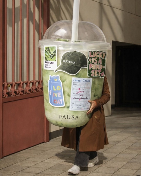 A person wears an oversized, realistic matcha drink cup costume featuring stickers and a guest check for a playful, experiential brand aesthetic.