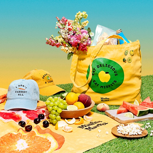 A high-conversion lifestyle creative featuring a branded grocery tote filled with fresh produce, flowers, and snacks on a sunny picnic setting.