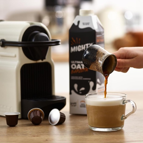 A high-quality lifestyle shot showing oat milk being poured from a small pitcher into a foamy latte, with the full product packaging and coffee machine in the background.