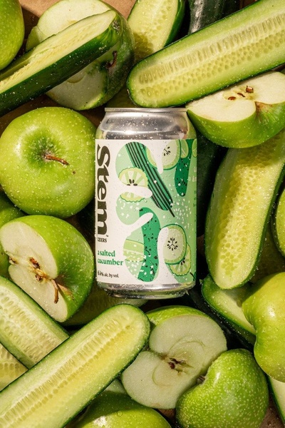 A high-angle, balanced still life shot featuring a can of salted cucumber cider surrounded by fresh, sliced green apples and cucumbers.