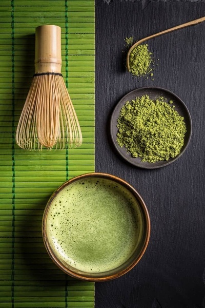 A high-contrast, overhead studio shot featuring fresh matcha powder, a bamboo whisk, and a prepared bowl of tea on a split green and black background.