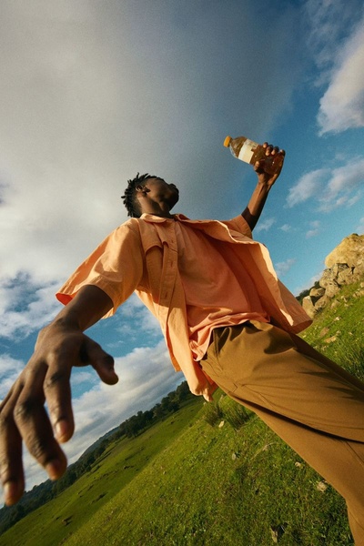 A low-angle, wide-shot photograph of a person outdoors in a field, holding a beverage bottle toward the sky to emphasize the product in an open, natural environment.