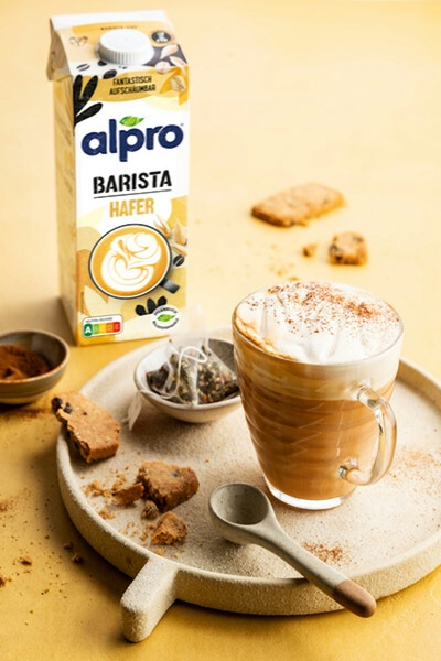 A high-quality lifestyle shot of Alpro Barista Oat Milk next to a freshly frothed cappuccino and cookies on a neutral textured plate.