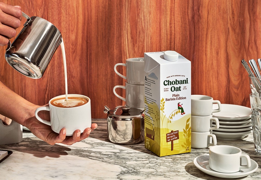 A high-quality lifestyle shot featuring a professional barista-style pour of oat milk into a coffee cup, highlighting the product packaging alongside the drink.