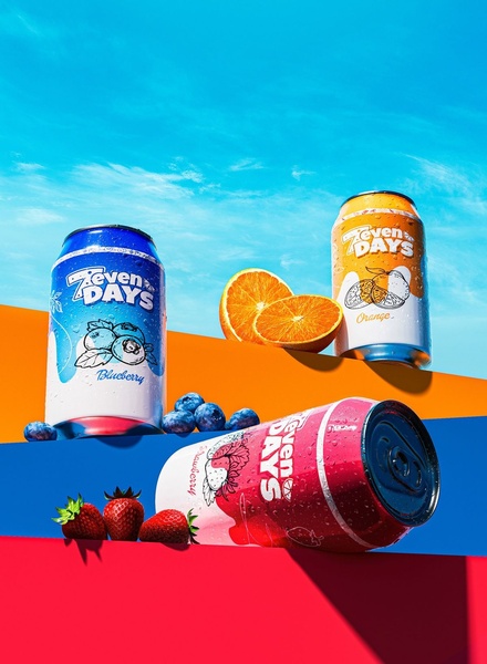 A visually striking product hero shot featuring blueberry, orange, and strawberry flavors of a 7-Days beverage set against a vibrant, multi-level colored background.