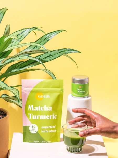 A high-key studio photograph featuring Matcha Turmeric packaging next to a glass of prepared drink, set against a bright yellow background with lush greenery.