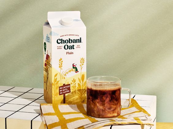 A high-quality, soft-lit product shot of an oat milk carton sitting on a checkered table with a freshly poured glass of coffee.