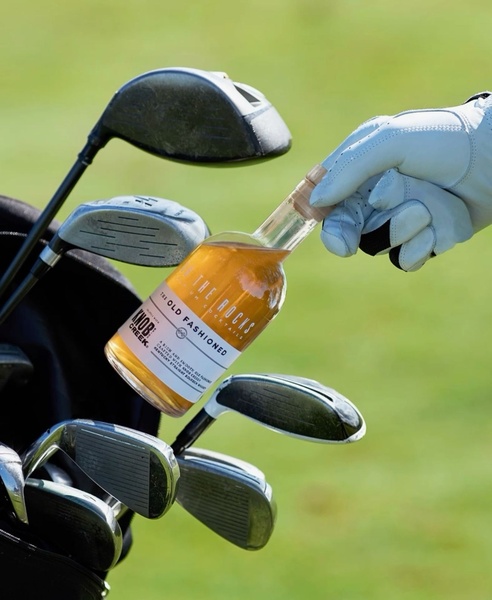 A high-end product shot featuring a cocktail bottle held near golf equipment, emphasizing a premium lifestyle and leisure.