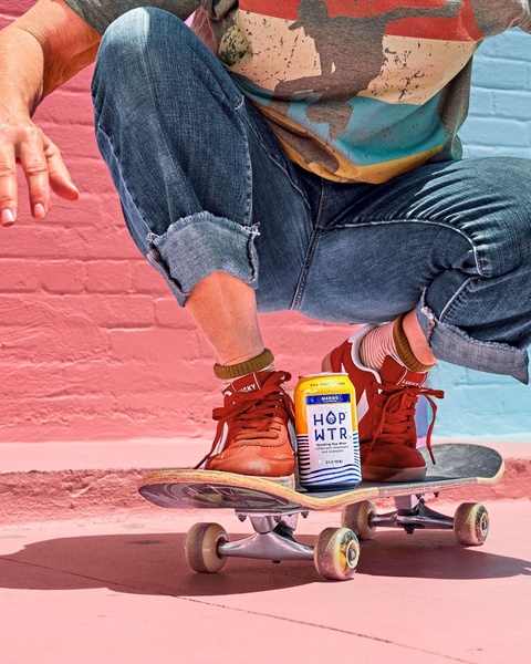 A close-up, 4:5 ratio shot of a skateboarder balancing a can of sparkling hop water on their board, set against a colorful, urban background.