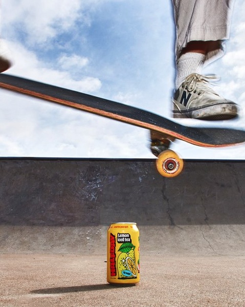 A high-energy, action-oriented shot featuring a lemon iced tea can centered on the ground with a skateboard jumping over it in mid-air.