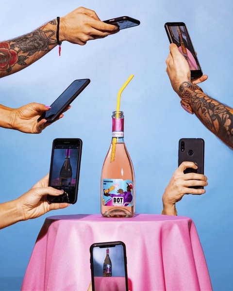A bottle of rose wine is placed in the center of a table, surrounded by multiple hands holding smartphones to capture photos of the product.