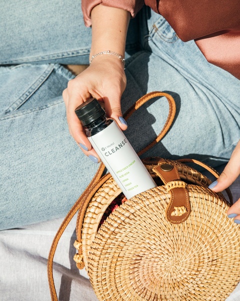 A close-up, natural-light shot featuring a person holding a wellness beverage bottle next to a stylish rattan bag, emphasizing a healthy lifestyle.