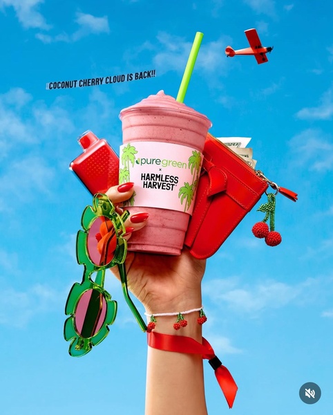 A high-energy, colorful product hero shot of a smoothie held against a bright blue sky, surrounded by summery props to signify a seasonal return.
