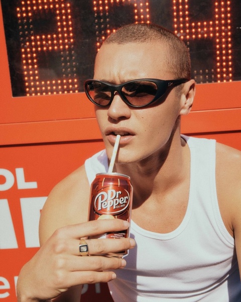 A stylish young man drinks a soda in front of a retro neon display. This visual uses a high-contrast lifestyle setting to anchor the product.
