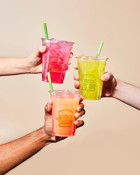 A close-up, high-energy shot of three hands toasting with colorful fruit-based beverages against a neutral studio background.