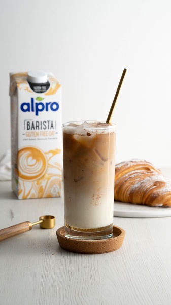 A high-quality lifestyle shot featuring a carton of barista-grade oat milk, an iced coffee beverage with a straw, and a croissant on a clean surface.