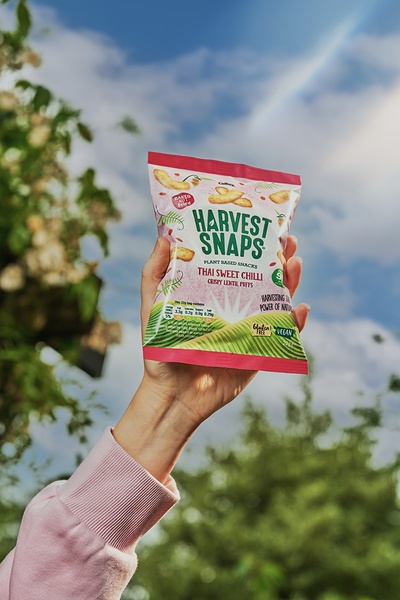 A high-angle, outdoor shot featuring a hand holding a bag of Thai Sweet Chilli lentil puffs against a bright, cloudy blue sky with natural lighting.