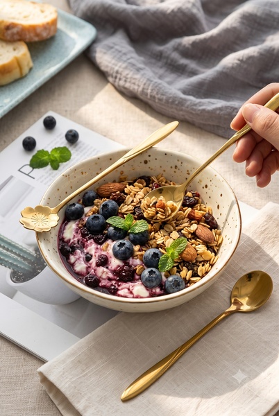 A top-down, brightly lit lifestyle shot of a bowl filled with granola, blueberries, and yogurt, featuring a gold flower-shaped spoon being lifted.