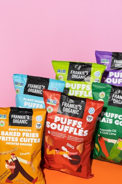 A high-energy, colorful product hero shot showcasing an assortment of organic plant-based snack bags against a soft pink background.