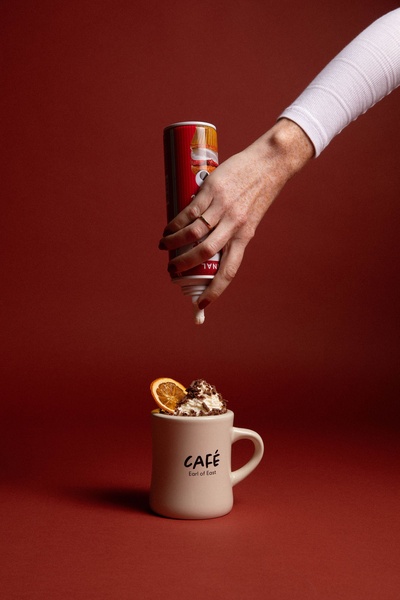 A close-up vertical shot showing whipped cream being dispensed onto a coffee mug topped with orange garnish against a dark red backdrop.