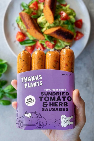 A high-quality, close-up product shot featuring plant-based sausages held in hand, with a finished meal blurred in the background to provide context.