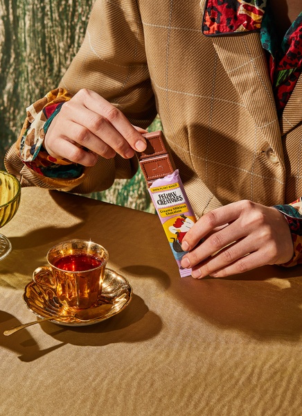 A high-end portrait featuring a chocolate bar held over a table with a gold-rimmed cup of tea, emphasizing a sophisticated, artisanal aesthetic.