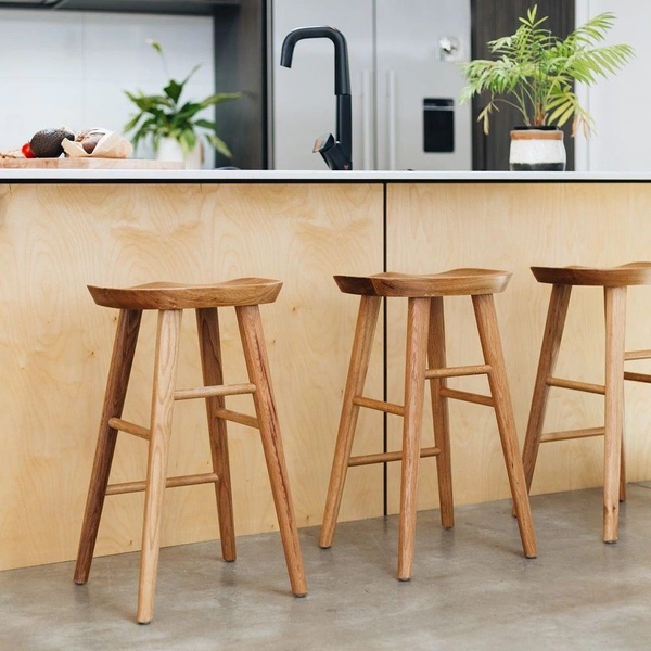 A high-quality 1:1 image showcasing wooden bar stools in a modern, light-filled kitchen setting with an emphasis on texture and natural materials.