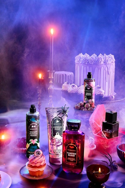 A dark, moody product staging featuring various body care items illuminated by candlelight and purple smoke, evoking a seasonal, atmospheric theme.