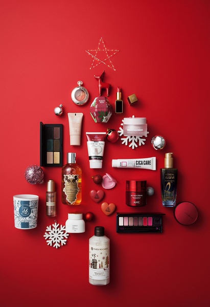 A high-angle flat lay arrangement of various beauty and skincare products organized into the shape of a Christmas tree against a bold red background.