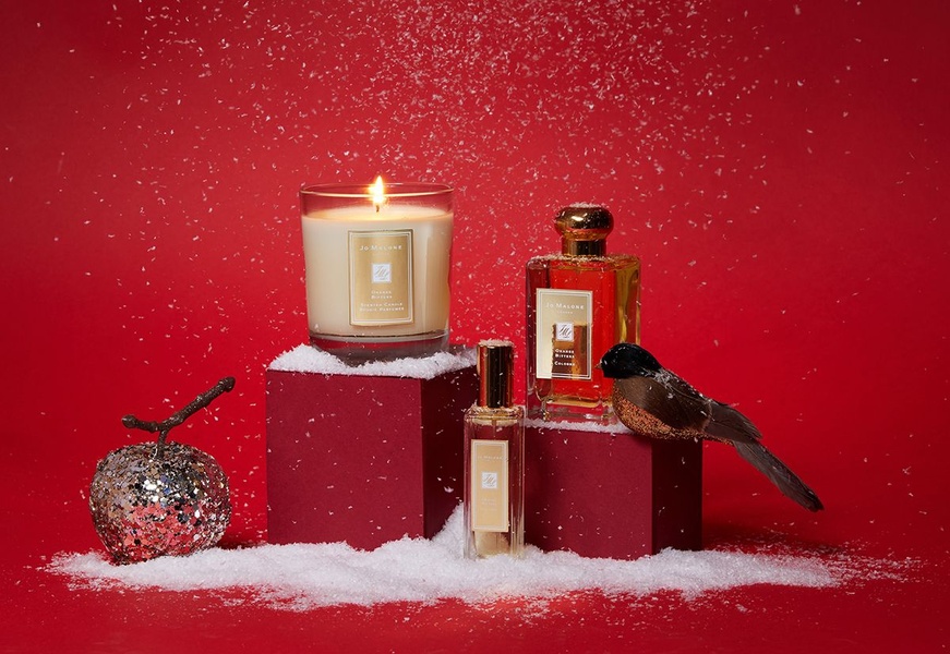 A high-end, seasonal product arrangement featuring a scented candle and cologne, showcased on elevated blocks amidst falling snow on a deep red background.