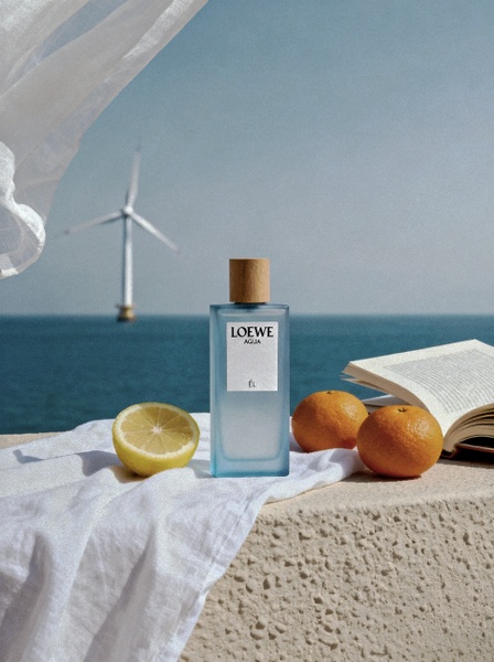 A high-end, bright, and minimalist product shot featuring a perfume bottle placed on a stone ledge by the ocean with fresh citrus.