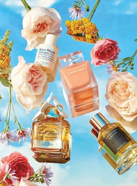 A high-aesthetic, top-down product shot featuring various perfume bottles floating amidst pastel flowers against a vibrant blue sky background.