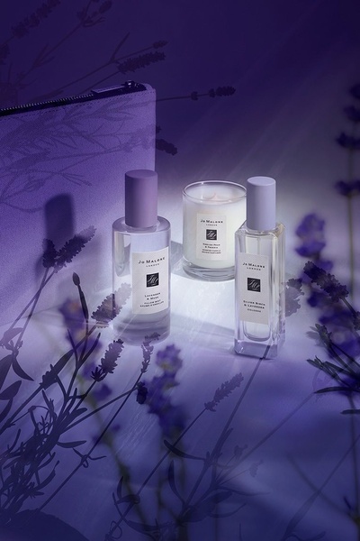 A high-end lifestyle product shot featuring a fragrance mist, a scented candle, and a perfume bottle arranged in a dimly lit, lavender-toned studio setting.