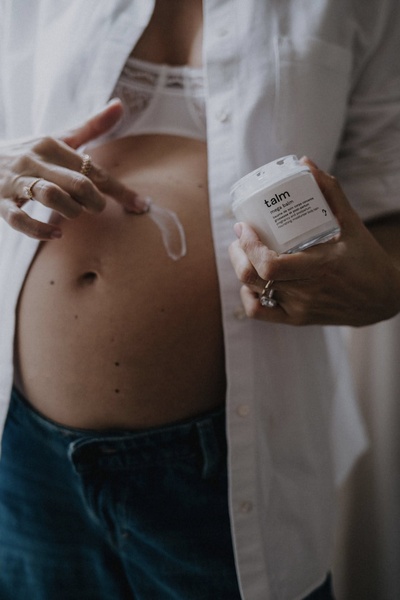 An intimate, close-up shot of a pregnant person applying a nourishing balm to their belly, highlighting the brand logo and product texture.
