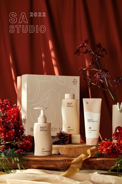 A high-end, seasonal display of three body care products arranged on a wooden surface with red curtains and dried foliage.
